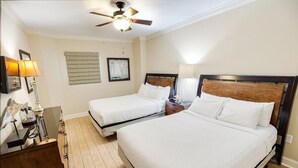 Quarto de hotel com duas camas, um ventilador de teto, uma escrivaninha com luminária e um quadro na parede.