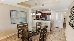 Uma sala de jantar com mesa e cadeiras de vidro, cozinha ao fundo e um quadro na parede.