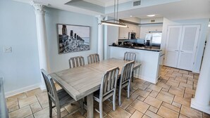 A dining area with a wooden table and chairs, a kitchen in the background, and a tiled floor. - Relax in Style: Oceanfront 3 Bedroom Condo (North Myrtle Beach)