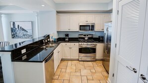 A modern kitchen with white cabinets, a black countertop, stainless steel appliances, and a tiled floor. - Relax in Style: Oceanfront 3 Bedroom Condo (North Myrtle Beach)
