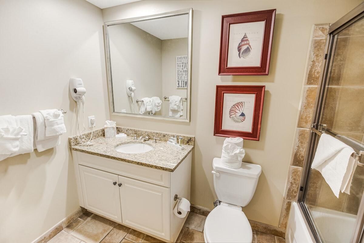 A bathroom with a sink, mirror, toilet, and shower. There are towels on the wall and a framed picture above the mirror.