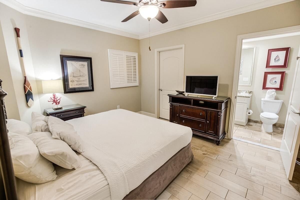 A bedroom with a bed, a dresser, a television, a ceiling fan, and a door leading to a bathroom.