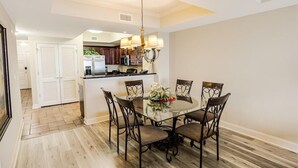 A dining area with a glass table and six chairs, a chandelier, and a kitchen in the background. - Coastal Comfort Redefined: 3 Bedroom Condo (North Myrtle Beach)