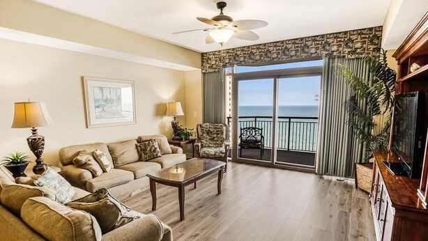 A spacious living room with a large sofa, a coffee table, and a chair. There is a balcony with a view of the ocean. - Coastal Comfort Redefined: 3 Bedroom Condo (North Myrtle Beach)