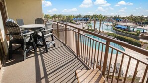 A balcony with a table and chairs, overlooking a pool area with umbrellas and palm trees. - Seaside Perfection 3 Bdrm Oceanfront w/ Whirlpool (North Myrtle Beach)