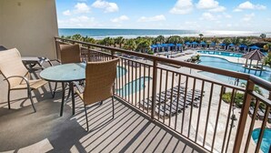 A balcony with a view of a pool and beach, furnished with wicker chairs and a table.