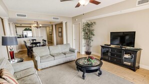 A living room with a grey sofa, a coffee table with a floral centerpiece, a TV stand, and a dining area in the background.