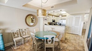 Un coin repas avec une table ronde et six chaises, une cuisine en arrière-plan et un luminaire décoratif au plafond.