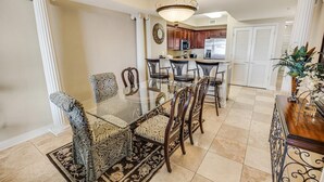 Un comedor con mesa de vidrio y sillas estampadas, una cocina al fondo y una planta decorativa a la derecha.