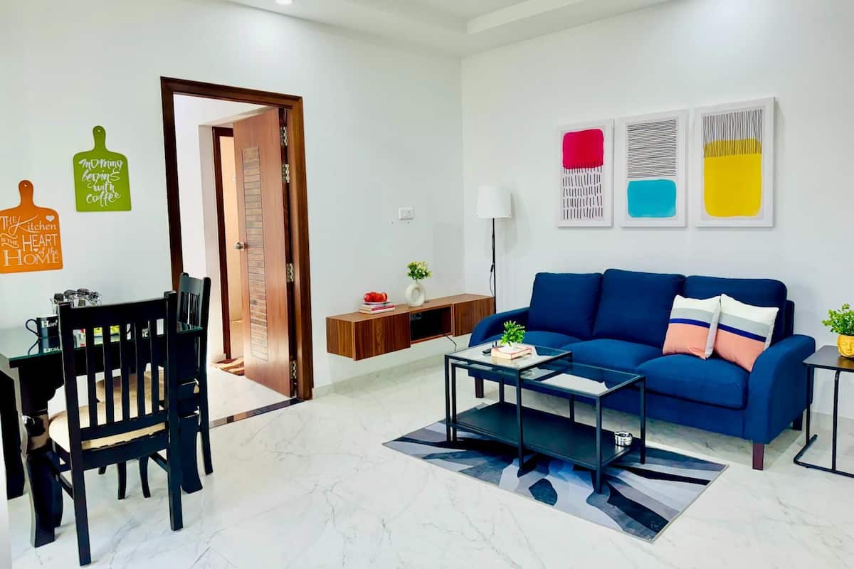 A modern living room with a blue sofa, a coffee table, a dining table with chairs, and a kitchen area visible through an open door.