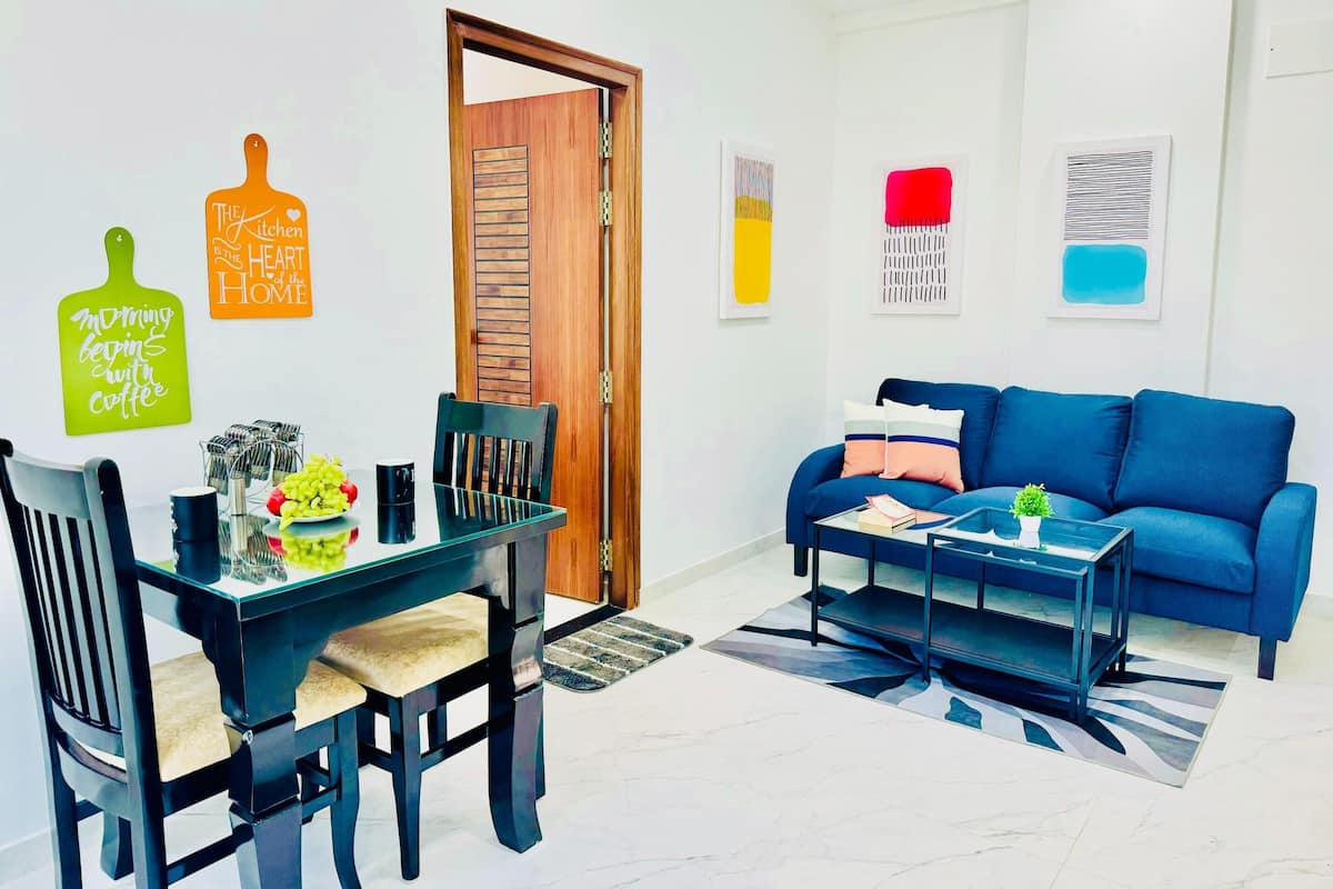 A modern living room with a dining area, featuring a blue sofa, a glass coffee table, and a dining table with chairs.