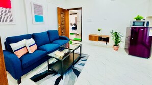 Una sala de estar moderna con un sofá azul, una mesa de centro de vidrio y una zona de cocina con refrigerador y microondas.