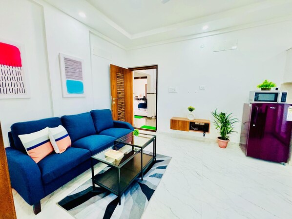 A modern living room with a blue sofa, a glass coffee table, and a kitchen area with a refrigerator and microwave.