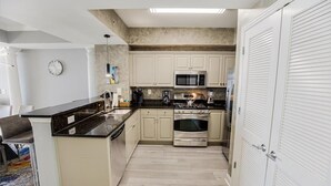 A compact kitchen with white cabinets, a black countertop, stainless steel appliances, and a clock on the wall. - 3 Bedroom Condo with King and Queen Sized Beds (North Myrtle Beach)