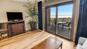 Un salon doté d’un téléviseur à écran plat, de meubles en bois, d’une grande plante en pot et d’un balcon offrant une vue sur la plage.