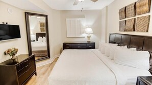 Une chambre à coucher avec un grand lit, une commode, un téléviseur, un ventilateur de plafond, un miroir et des œuvres d’art décoratives au mur.