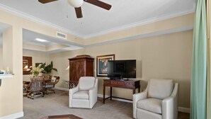 Una sala de estar con dos sillones blancos, una mesa de centro de madera, un televisor sobre un mueble bajo, un comedor con sillas de mimbre y una planta grande.