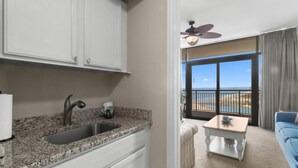 A kitchen with a granite countertop, a sink, and white cabinets. There is a balcony with a view of the beach and a ceiling fan. - Relax and Unwind in this Oceanfront 1 Bdrm Condo (North Myrtle Beach)