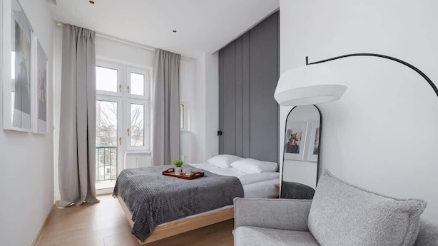 Un dormitorio moderno con cama, sofá gris, mesita de noche, espejo y ventana con cortinas.