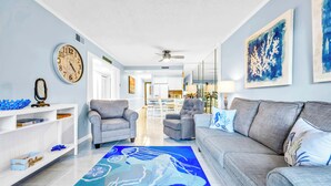 Ein modernes Wohnzimmer mit einem grauen Sofa, zwei Sesseln, einem Couchtisch und einem blauen Teppich mit Muster. An den Wänden hängen Wanduhren und gerahmte Kunstwerke.