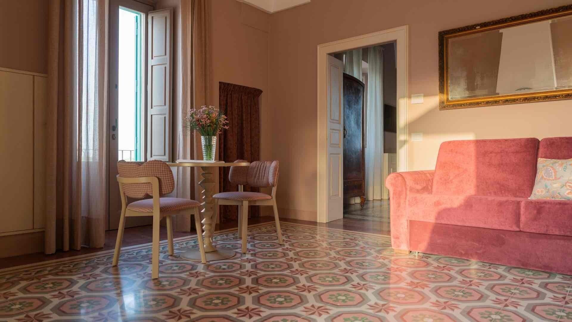 A room with a patterned floor, a pink sofa, a dining table with chairs, and a vase of flowers.
