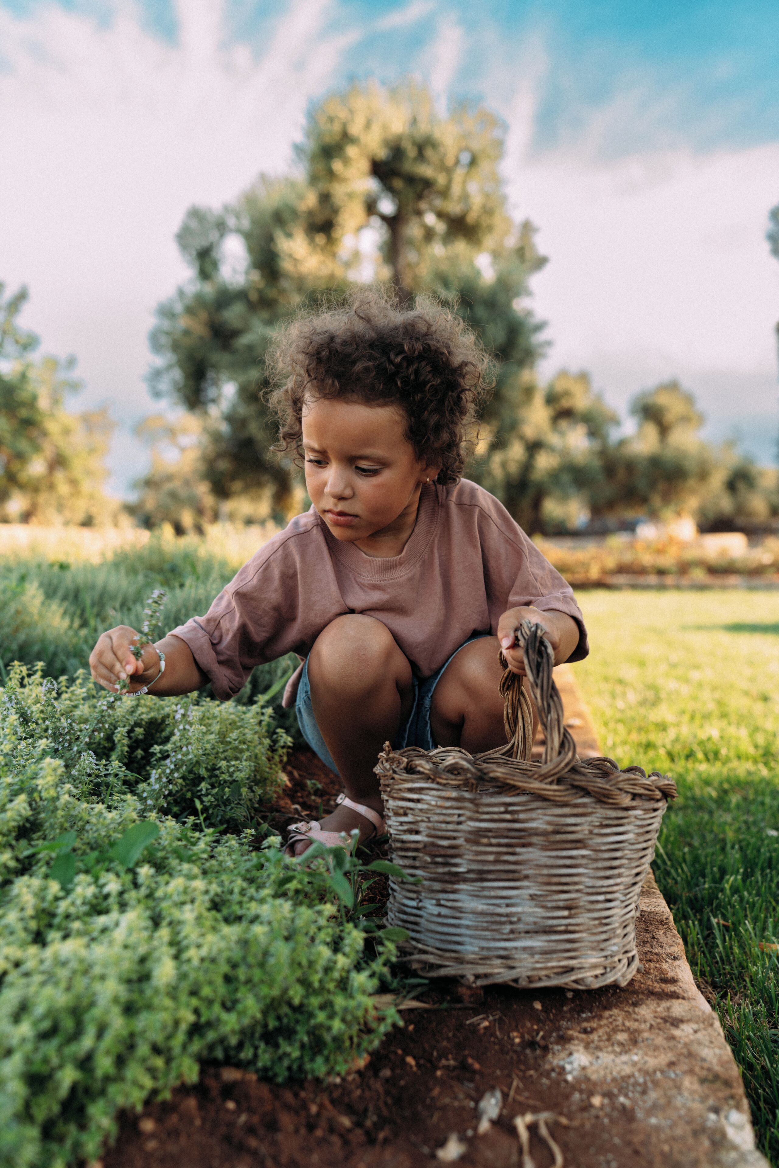 A child crouching on the ground near a basket, surrounded by greenery.