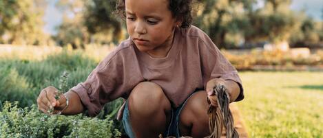 A child crouching on the ground near a basket, surrounded by greenery.