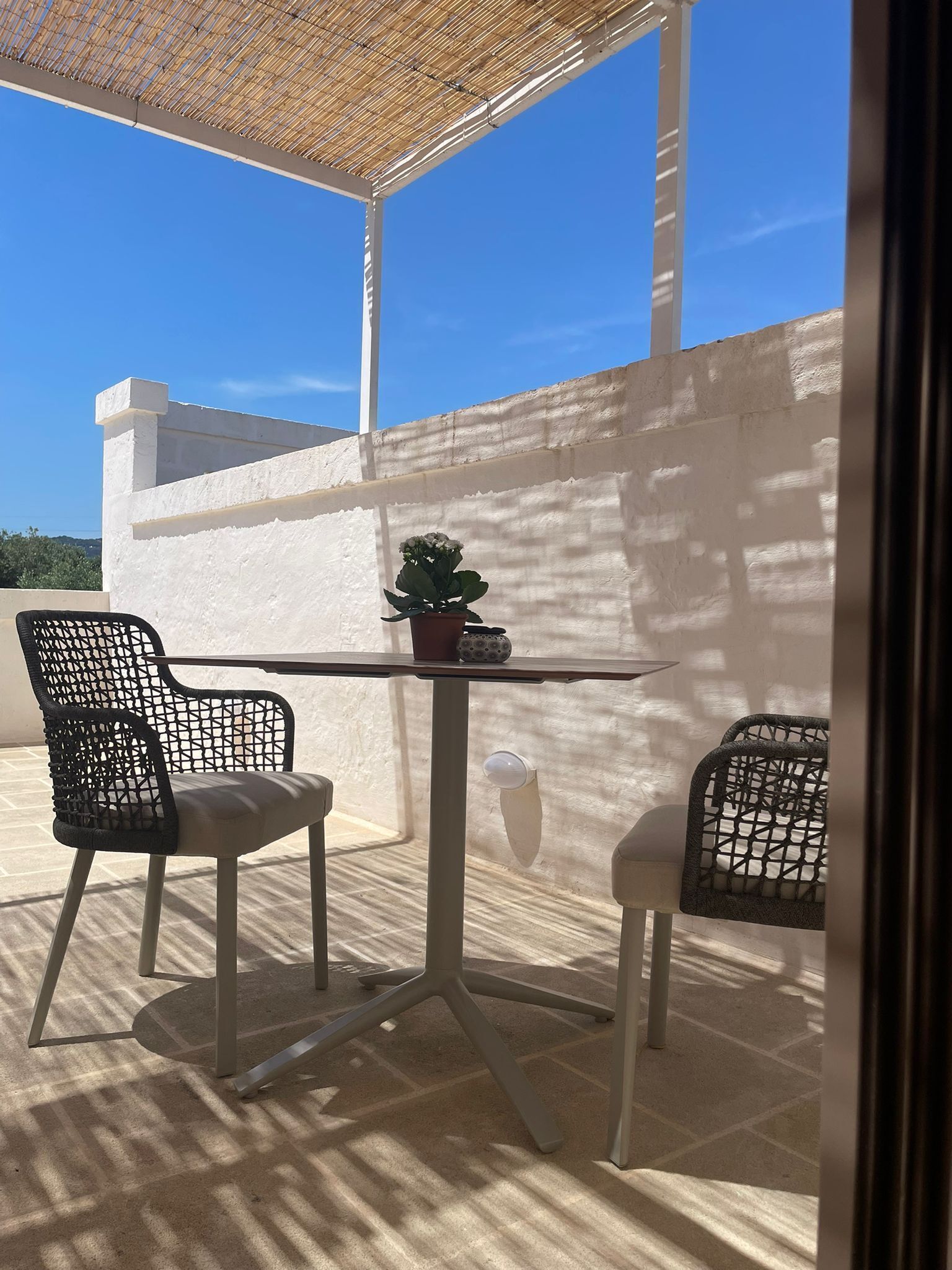 A patio with two chairs and a table, a potted plant, and a white wall.