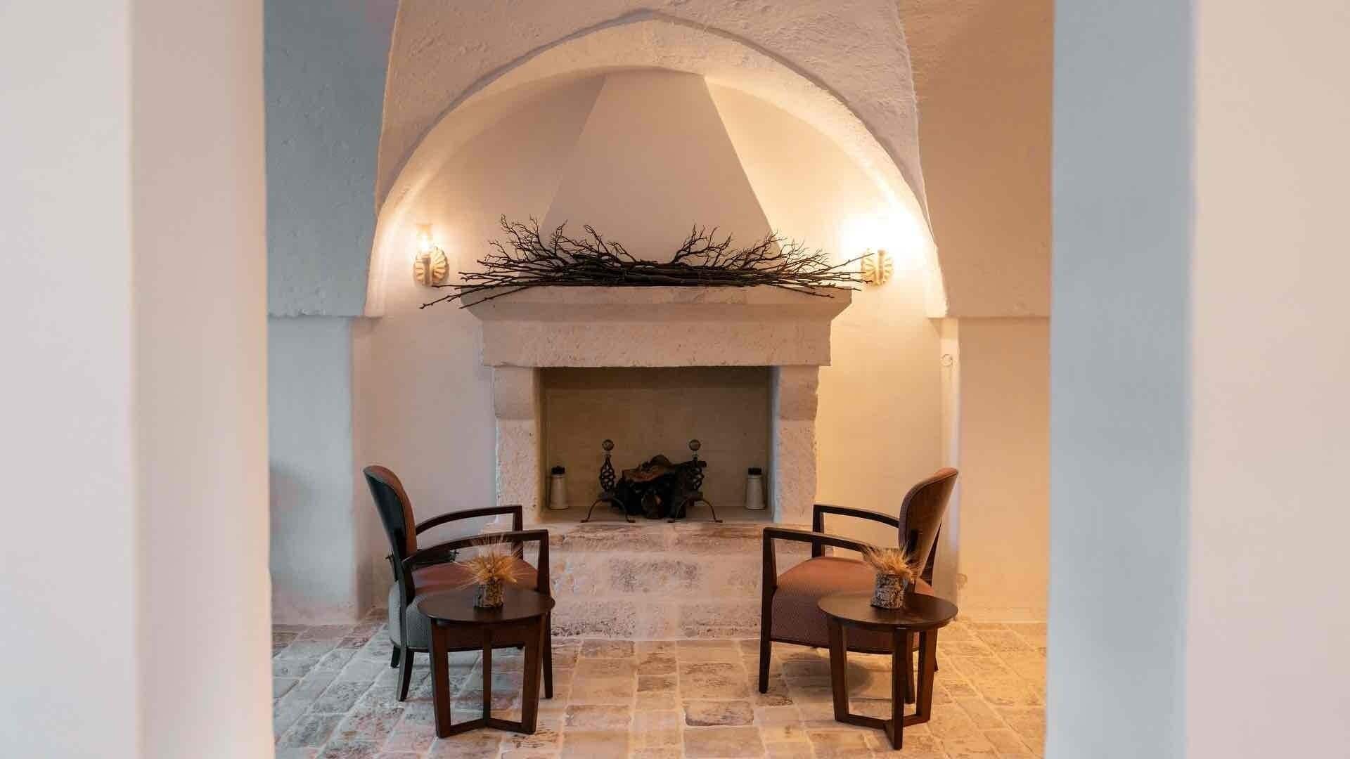 Un coin chaleureux avec une cheminée, deux fauteuils placés face au foyer, un sol en pierre et une branche décorative au-dessus du feu.