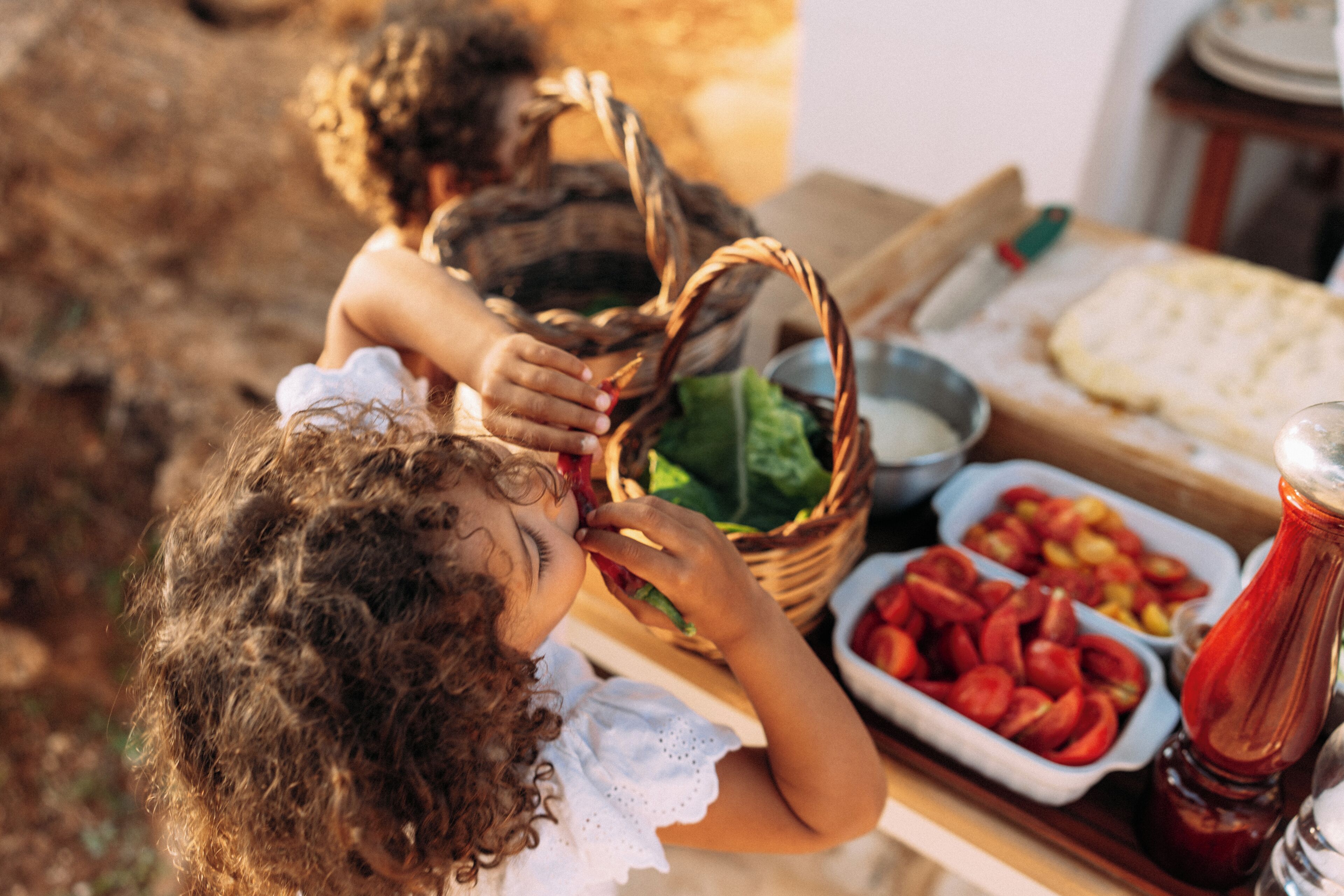 A child eating an apple at a picnic table with a basket, a bowl of sliced tomatoes, and a plate of bread.