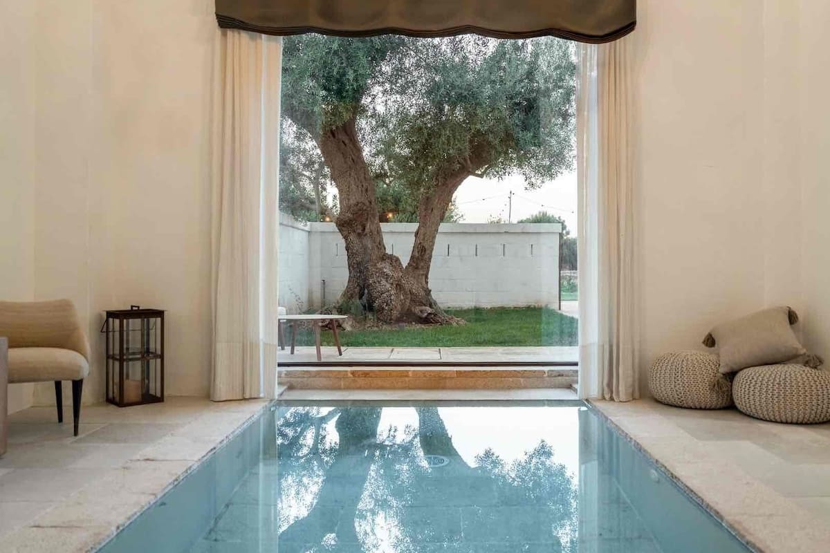 Ein Zimmer mit Pool, einem Sessel, einem kleinen Tisch und Blick auf einen Baum und ein Gebäude.
