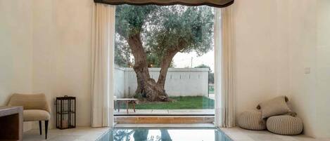 A room with a pool, a chair, a small table, and a view of a tree and a building outside.