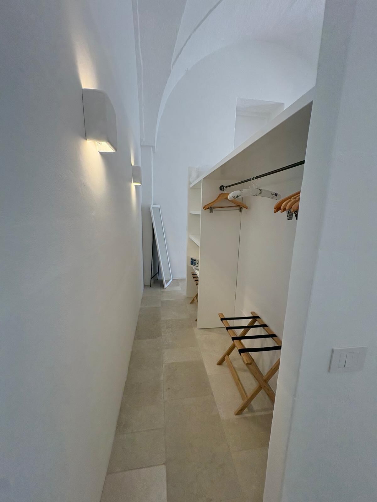 A narrow hallway with white walls and a tiled floor, leading to a closet area with hanging clothes and a wooden bench.