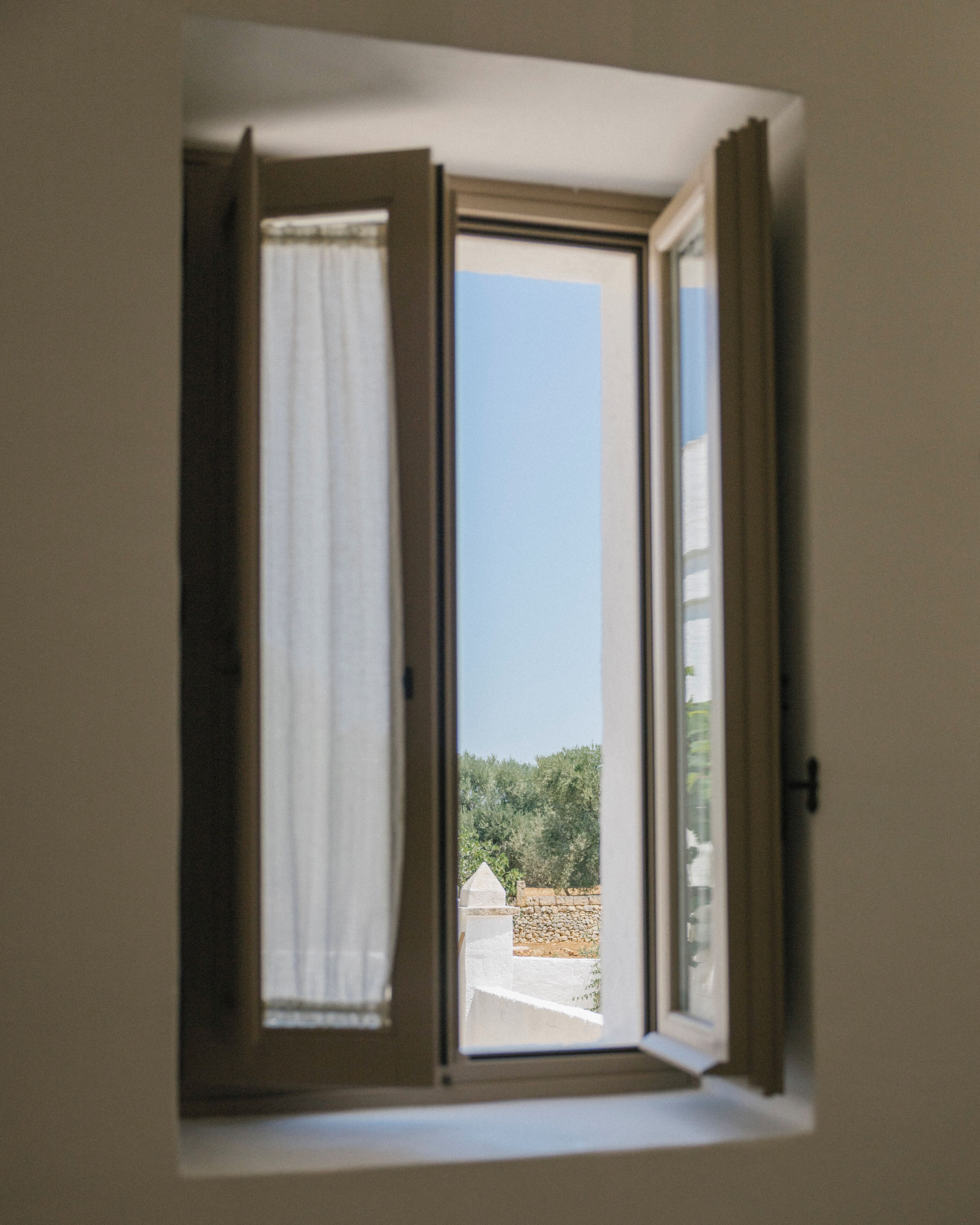 A window with an open curtain, a clear view of the sky, and a stone wall with greenery in the background.