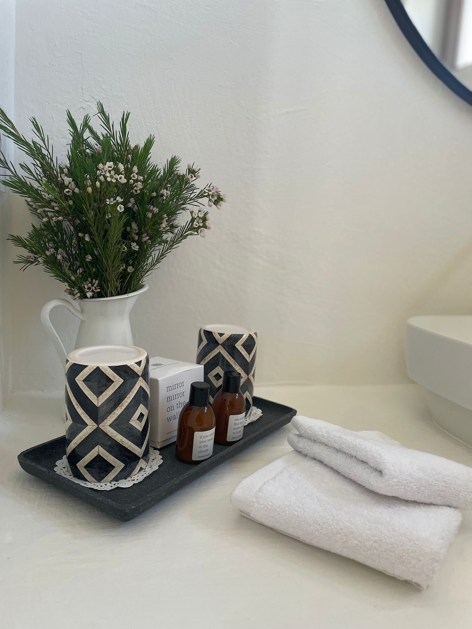 A bathroom with a white sink, a white towel, a black tray with a white mug, a black and white mug, a white pitcher with green foliage, and two small bottles.