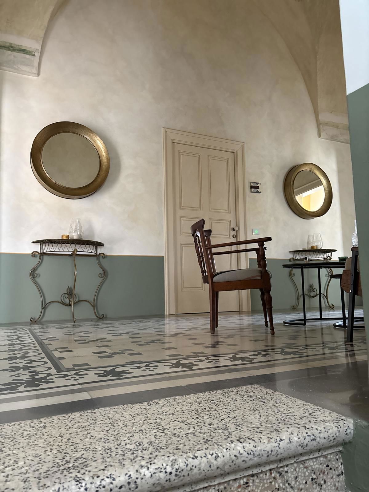 Una habitación con un amplio arco de entrada, dos espejos redondos, una silla de madera, una mesita con un vaso y una alfombra estampada.