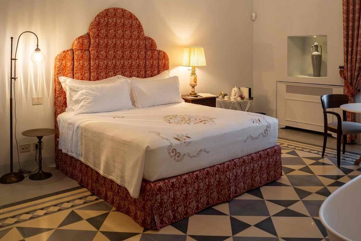 Ein Hotelzimmer mit geblümter Bettdecke, einer Nachttischlampe, einem Schreibtisch mit Stuhl und einem gemusterten Boden.
