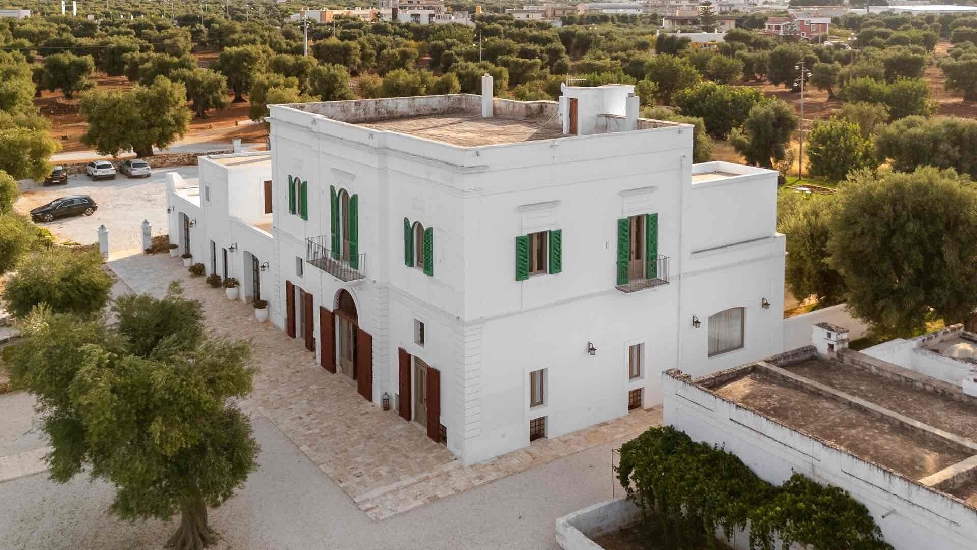Un edificio bianco con persiane verdi, un cortile interno e alberi di ulivo circostanti.