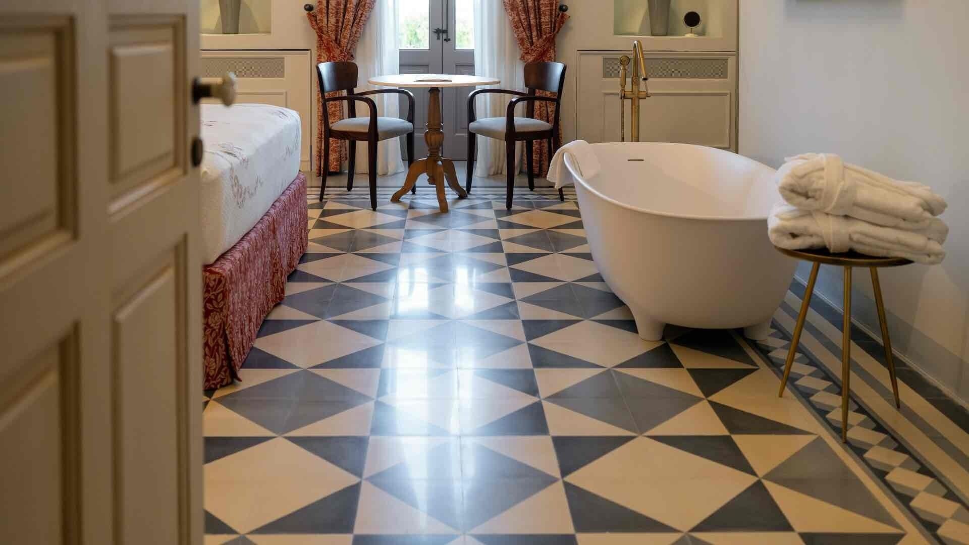 Une salle de bain avec un sol à motifs géométriques, une baignoire, une table ronde avec des chaises et un lit recouvert d’une couvre-lit à motifs.