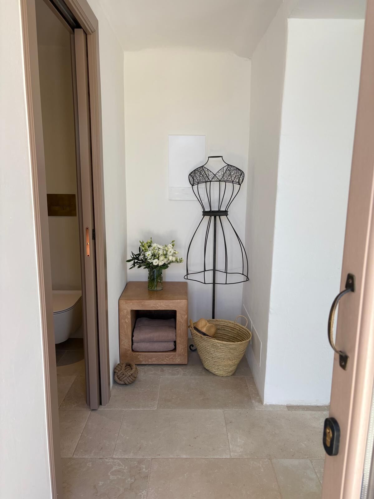 A hallway with a mannequin dress form, a small table with a vase of flowers, a woven basket, and a door.