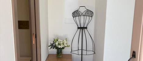 Un pasillo con un maniquí para vestidos, una mesita con un jarrón de flores, una cesta tejida y una puerta.