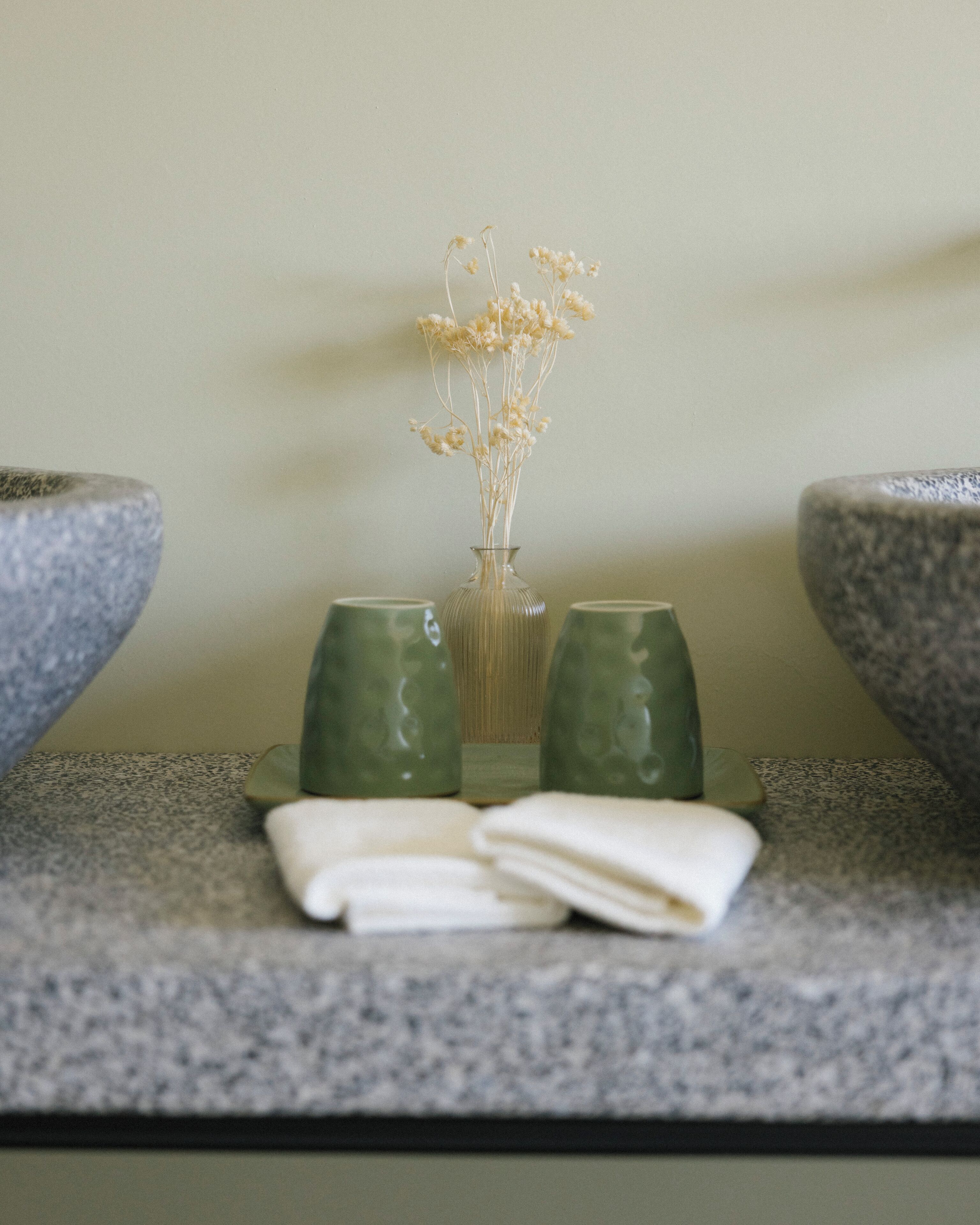 Ein Badezimmer-Waschtisch mit einer Vase getrockneter Blumen, zwei grünen Gläsern und einem gefalteten Handtuch.