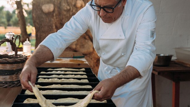 A chef preparing food outdoors, with a table and a large tree in the background.
