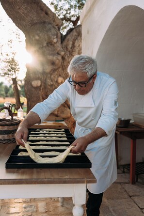 Un chef préparant des plats à l’extérieur, avec une table et un grand arbre en arrière-plan.