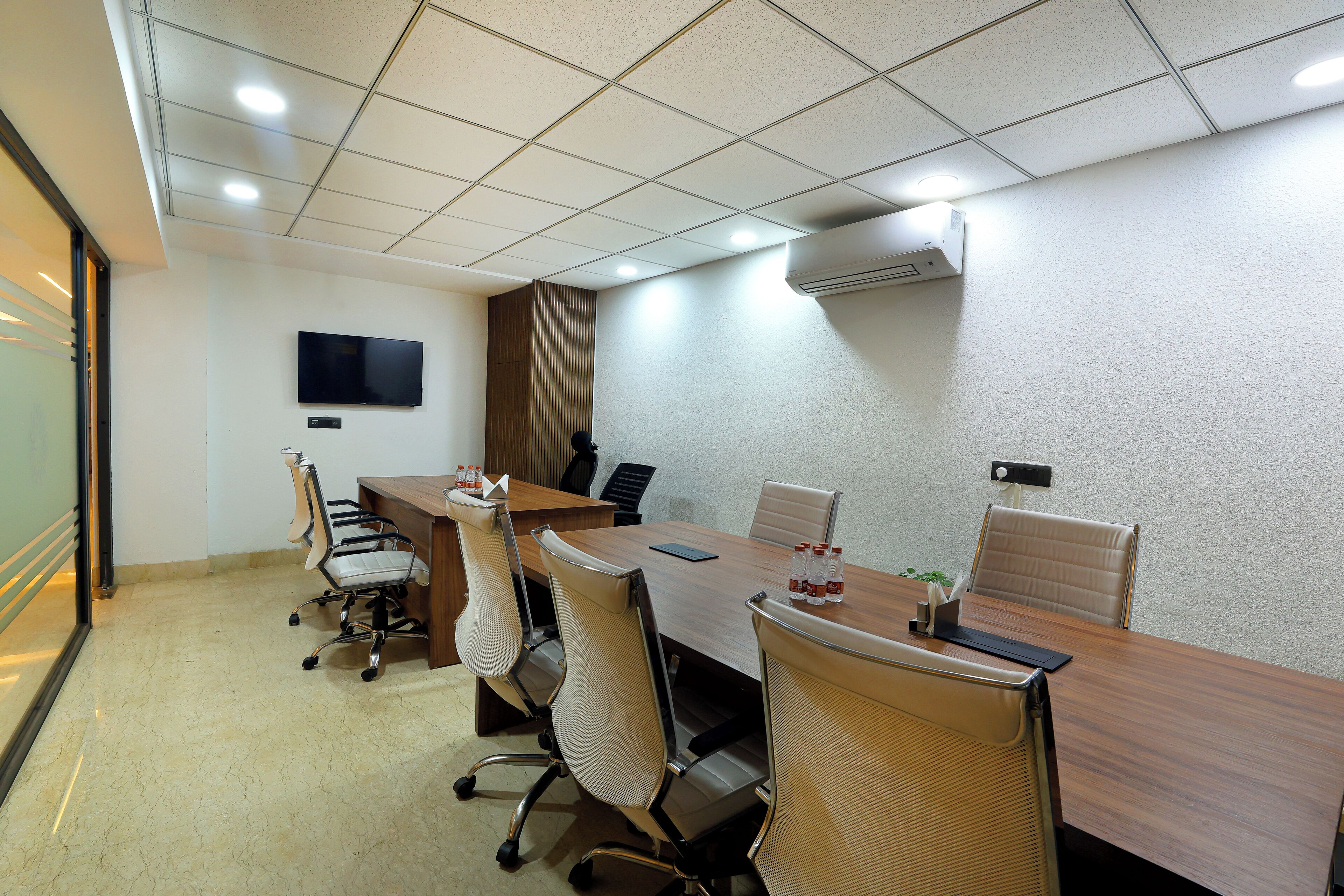 A conference room with a long table, six chairs, a flat-screen TV, and an air conditioning unit on the wall.
