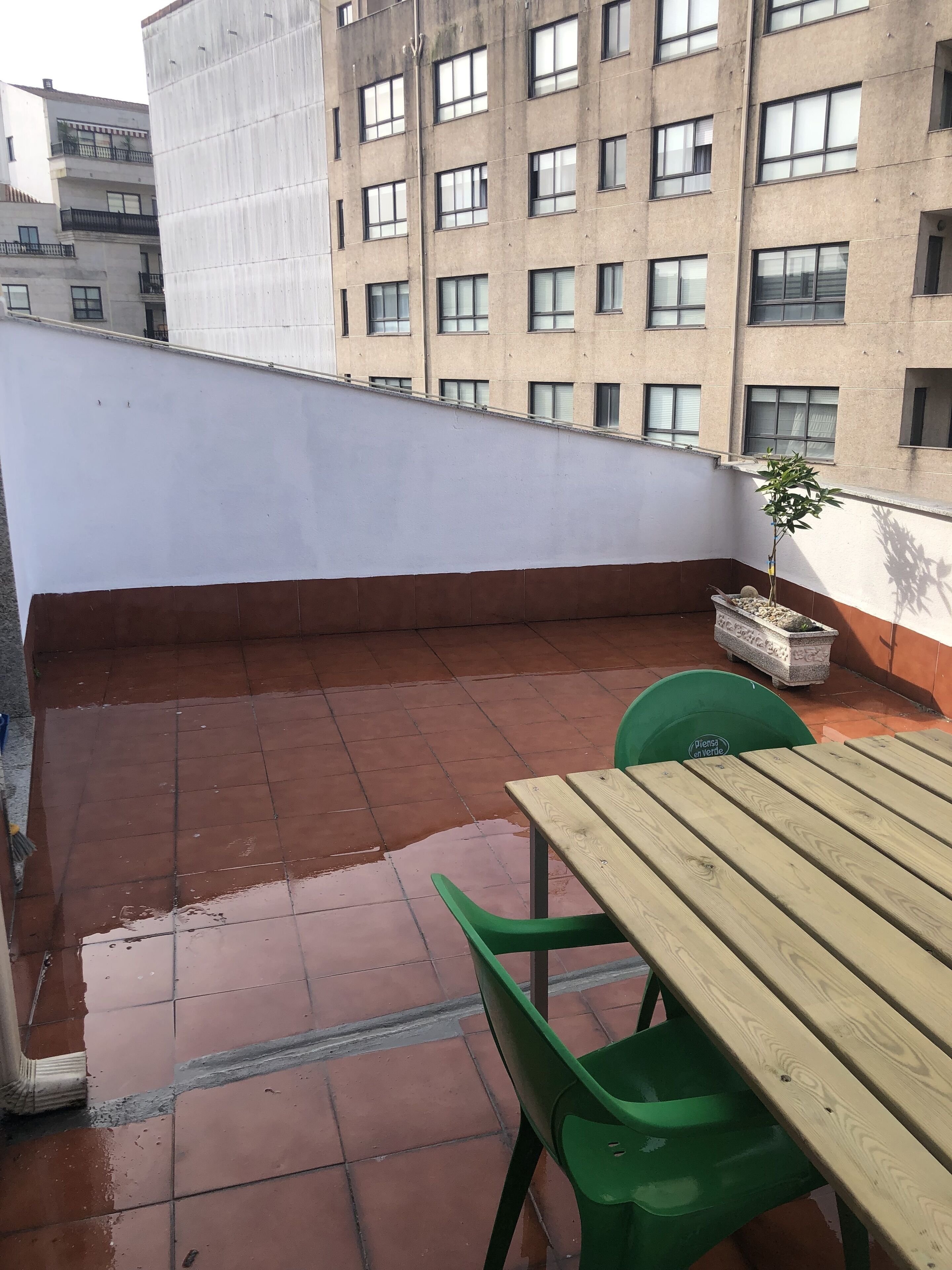 Um terraço no telhado com mesa de madeira e cadeiras verdes, uma planta em vaso e vista de um edifício de vários andares.