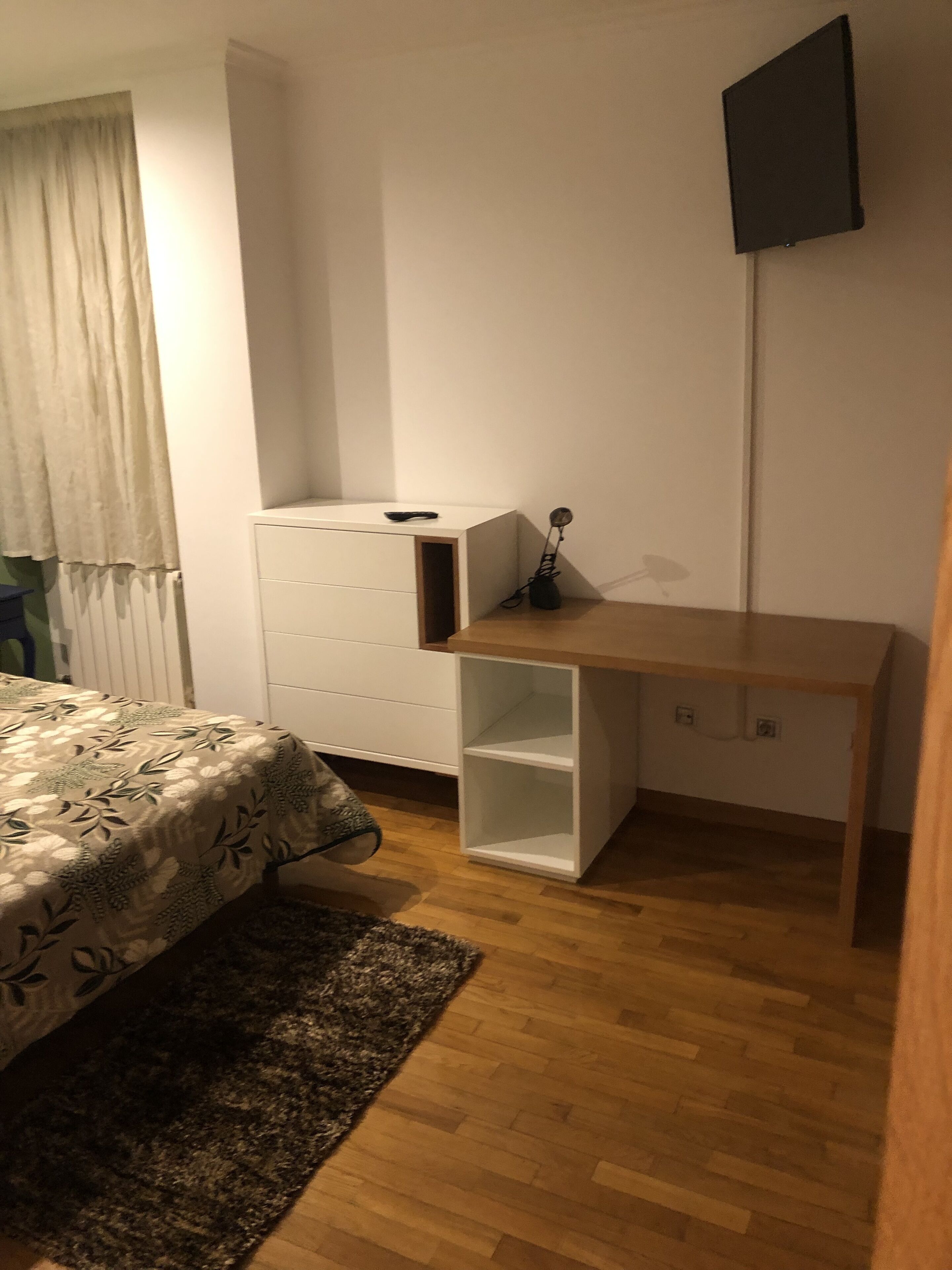 Una camera da letto con un letto, una cassettiera bianca, una scrivania in legno, una televisione a parete e una finestra con tende.