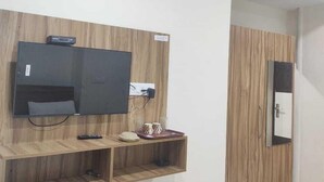 A room with a TV mounted on a wooden panel, a small shelf with a remote control, and a wardrobe with a mirror. - Tirath Hotel - Rishikesh - Uttrakhand (Rishikesh)