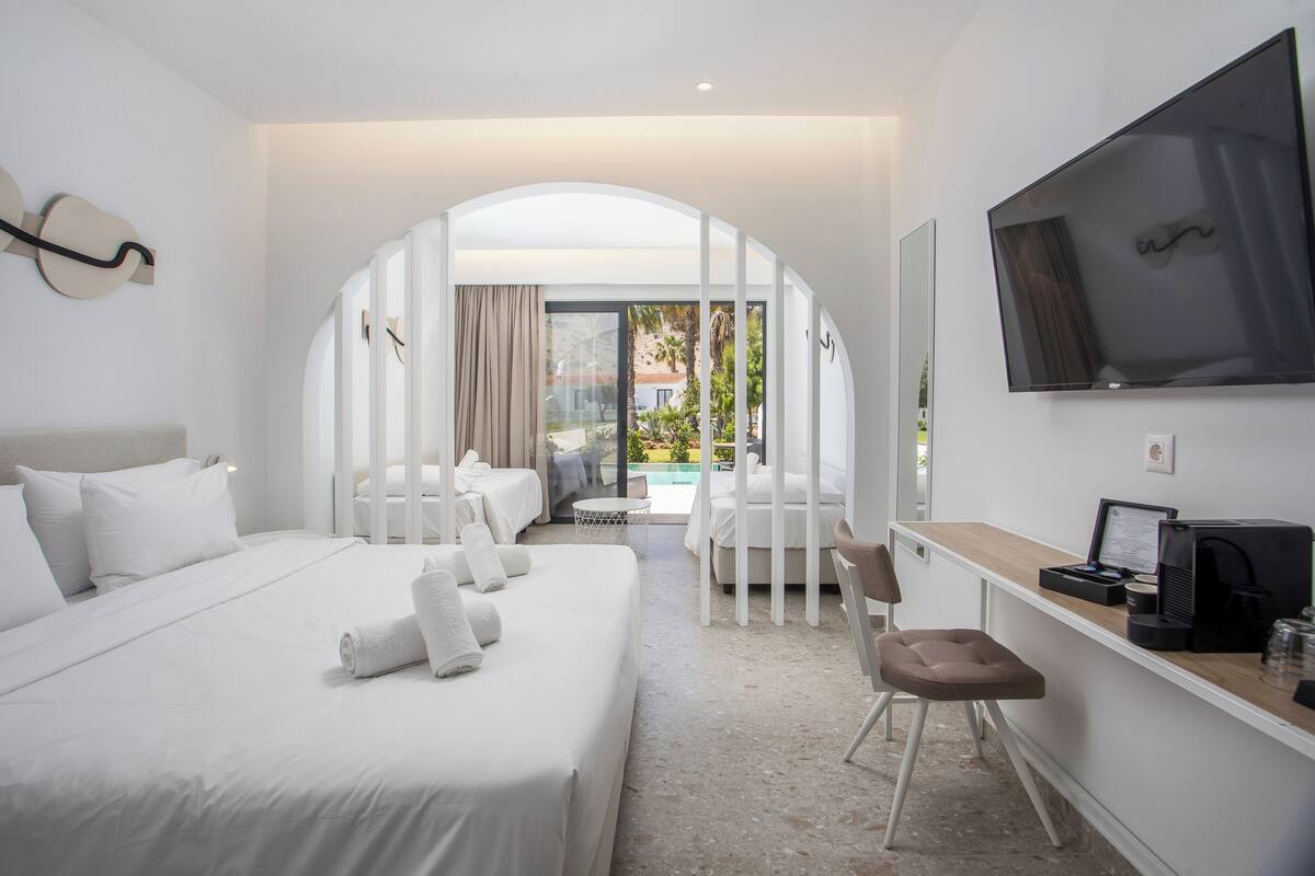 A modern hotel room with a large bed, a desk with a computer, a TV, and a view of the outdoor pool area.