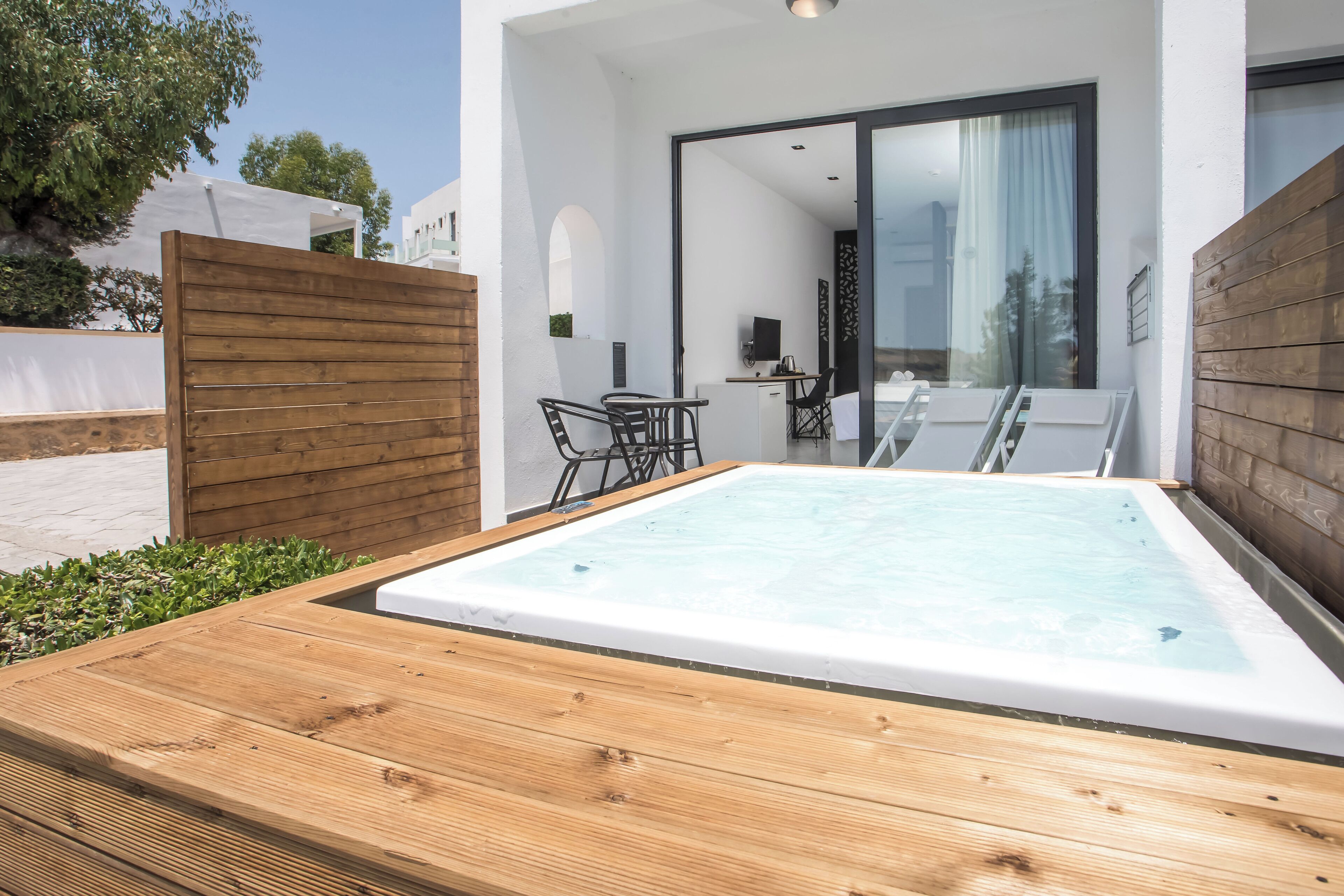 A modern outdoor pool area with a wooden deck, a seating area with chairs, and a glass door leading to a room.